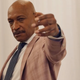 Actor Tony Todd sports a bald head and mustache and is wearing a dark tie and tan suit while gesturing toward the camera in a scene from Shudder's Horror Noire.